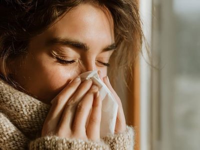 Young woman with cold or flu blowing nose into tissue while wearing cozy knit scarf near window with natural light.